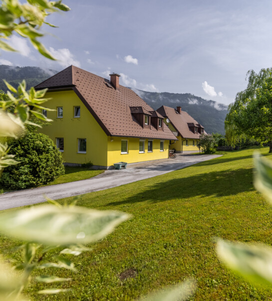 Außenansicht der Wohngemeinschaft, neben einer kleinen Einfahrt. In der Umgebung sind Bäume, Büsche und eine große Wiese.