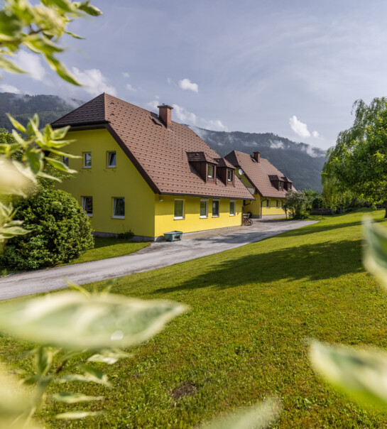 Außenansicht der Wohngemeinschaft, neben einer kleinen Einfahrt. In der Umgebung sind Bäume, Büsche und eine große Wiese.