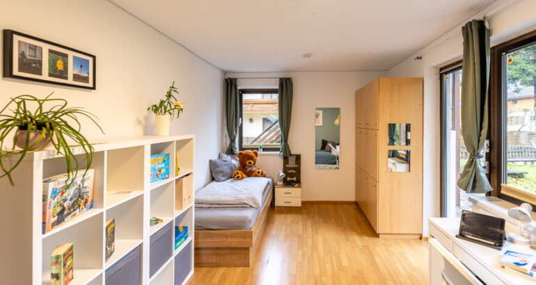 Innenansicht eines Schlafzimmers der Wohngemeinschaft mit Bett, Regalen und Schreibtisch. Fenster erlauben viel Licht in das Zimmer.