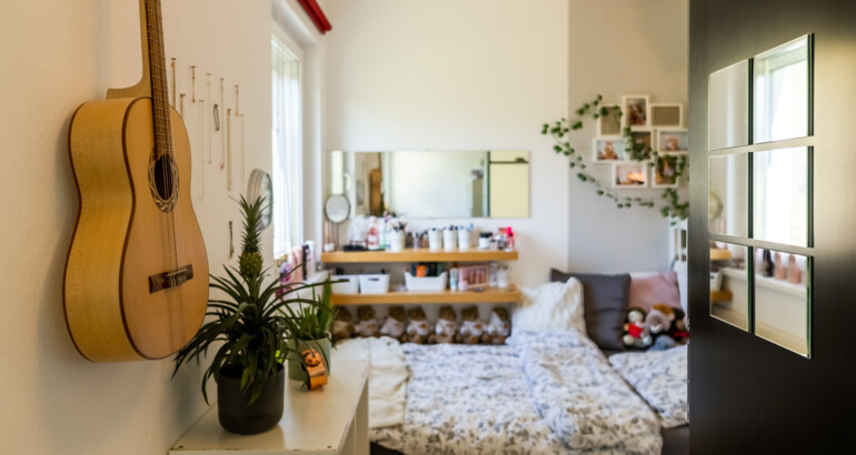 Innenansicht eines Schlafzimmers der Wohngemeinschaft mit Bett im Hintergrund und einer Gitarre die an der Wand hängt im Vordergrund.