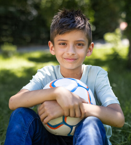 Bub sitzt in der Wiese, mit einem Ball in der Hand und lächelt in die Kamera.