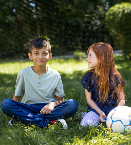 Zwei Kinder sitzen in der Wiese, neben ihnen liegt ein Fußball.